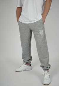 Gray sweatpants with elastic cuffs, featuring a small logo in light gray. Worn with a white t-shirt and white sneakers.