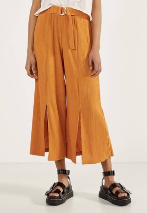 Person trägt orangefarbene, weit geschnittene, gekürzte Hose mit vorderen Schlitzen und schwarze Plateau-Sandalen mit Schnallen vor hellem Hintergrund.