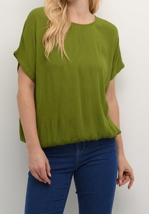 Femme portant un haut ample à manches courtes vert olive avec un col rond et un jean bleu ajusté sur un fond blanc uni.