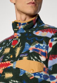 Man in een kleurrijke fleece pullover met vissenpatroon, voorzien van een beige drukknoop borstzak en het Patagonia-logo op de borst.
