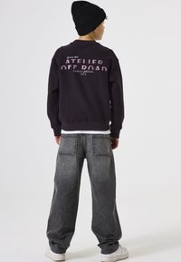 Sudadera morada oscura de corte oversize con texto impreso en la parte posterior; combinada con jeans grises holgados y zapatillas blancas. Gorro negro en la cabeza.