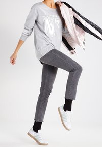 Šedá mikina s potiskem písmen, šedé skinny džínsy, bílé tenisky se zelenými pruhy a světle růžová prošívaná bunda přehozená přes rameno.