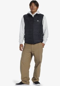 Black padded vest over a gray hoodie, featuring a front zipper and logo. Tan pants and black shoes complete the outfit.
