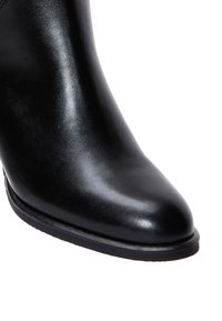 Bota de tobillo de cuero negro que presenta una textura suave, punta afilada y un tacón delgado. La suela es plana y ligeramente texturizada.