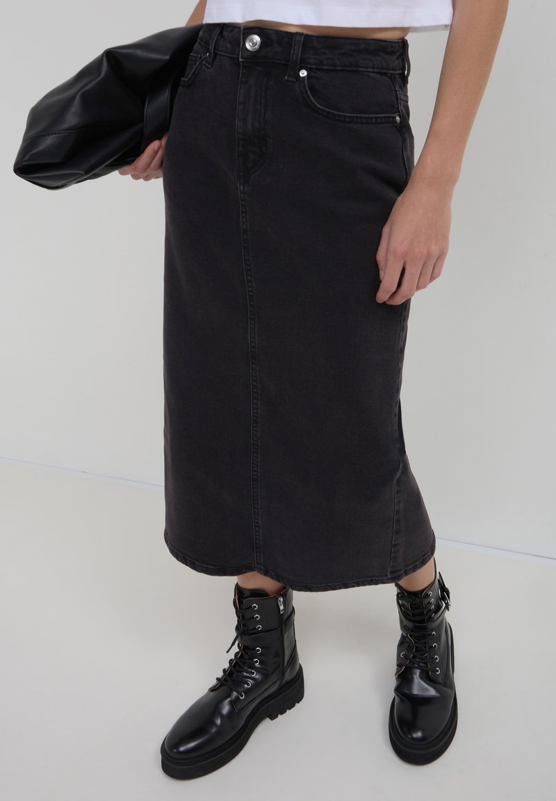Donna che indossa una gonna midi di jeans nera, stivali da combattimento neri con lacci, mentre tiene in mano una giacca di pelle nera, su uno sfondo chiaro e uniforme.