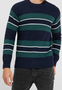 Pull en maille bleu marine avec des rayures horizontales en vert foncé, vert clair et blanc. Col rond et poignets et ourlet côtelés.