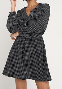 Schwarzes Kleid mit weißen Polka-Dots, Rüschenausschnitt, langen Ärmeln, Bindegürtel und ausgestelltem Rock, aus leichtem Stoff gefertigt.