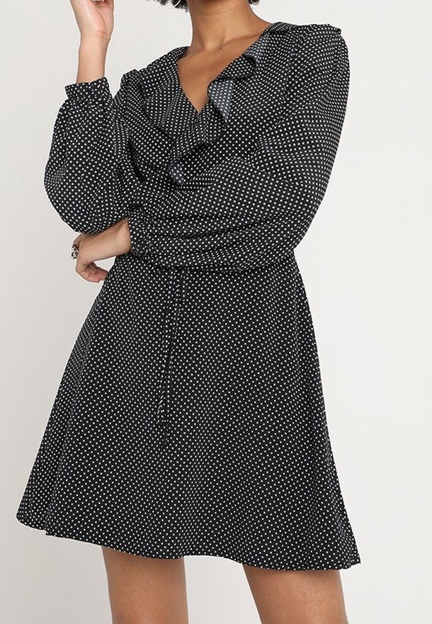 Schwarzes Kleid mit weißen Polka-Dots, Rüschenausschnitt, langen Ärmeln, Bindegürtel und ausgestelltem Rock, aus leichtem Stoff gefertigt.