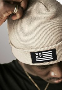 Tan knitted beanie with a ribbed texture and a rectangular black patch featuring white stripes and symbols. Close-up, showing hand adjusting the hat.