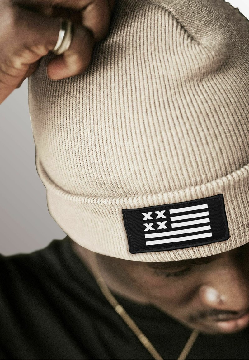 Tan knitted beanie with a ribbed texture and a rectangular black patch featuring white stripes and symbols. Close-up, showing hand adjusting the hat.