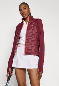 Giacca a vento bordeaux con zip arancione, maniche lunghe e un design aderente, indossata sopra una camicia bianca e una gonna, tenuta con una racchetta da tennis.