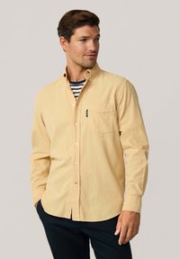 Camisa de botón color amarillo claro con un bolsillo en el pecho, diseño con cuello y mangas largas. Lleva puesta una camiseta de cuello redondo a rayas debajo.