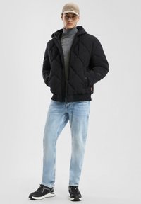Giacca a vento nera trapuntata con cappuccio, chiusura con zip e polsini elasticizzati, abbinata a jeans azzurri chiari e scarpe sportive nere. Cappello beige in testa.