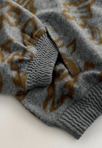 Grå fleece beklædning med et brunt abstrakt mønster. Har ribbede manchetter og en sidelomme med glat teksturdetaljering.