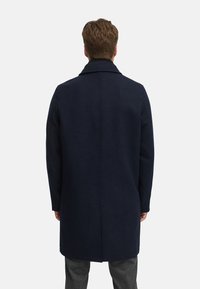 Navy Wolle Übermantel mit hohem Kragen, geradem Schnitt und subtilen Nahtdetails. Der Stoff hat eine glatte Textur und eine saubere Verarbeitung.