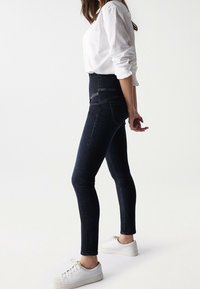 Žena v tmavých vysokých skinny džínách, bílé košili s ohrnutými rukávy a bílých teniskách, stojící bokem na jednoduchém pozadí.