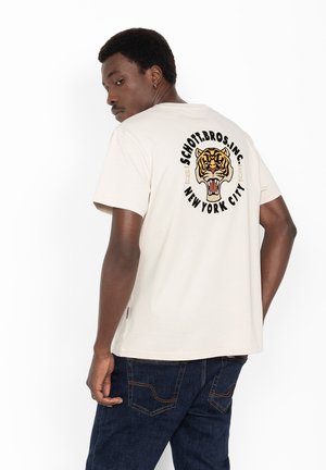 Mężczyzna w beżowej koszulce z ryczącym tygrysem i napisem "Schott Bros. Inc. New York City" na plecach, w ciemnoniebieskich dżinsach.