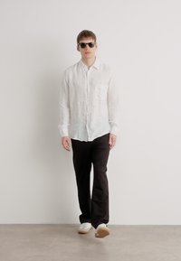 Camisa blanca de lino de botones con un bolsillo, combinada con pantalones negros y zapatillas blancas. El modelo lleva gafas de sol oscuras y está de pie sobre una superficie clara.