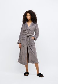 Pilkos spalvos vilnonis chalatas su ilgomis rankovėmis, diržu, dviem priekinėmis kišenėmis ir purus tekstūra. Dėvimas su juodais šlepetėmis.