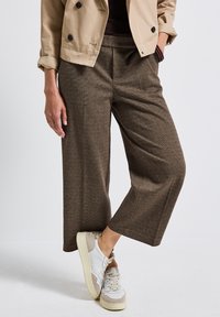Person med korta bruna vida byxor, en beige dubbelknäppt jacka med uppvikt ärm, och vita sneakers som står mot en enfärgad bakgrund.