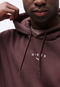 Κοντινό πλάνο ενός ατόμου που ρυθμίζει τη κουκούλα μιας μαroonς φούτερ με κουκούλα, στην οποία είναι κεντημένο το "HYROX" και ένα μικρό λογότυπο Puma στο στήθος.