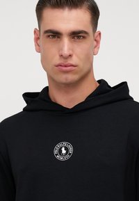 Felpa nera con cappuccio in tessuto morbido, dotata di cordino, con un logo bianco circolare sul petto. Design semplice ed elegante.