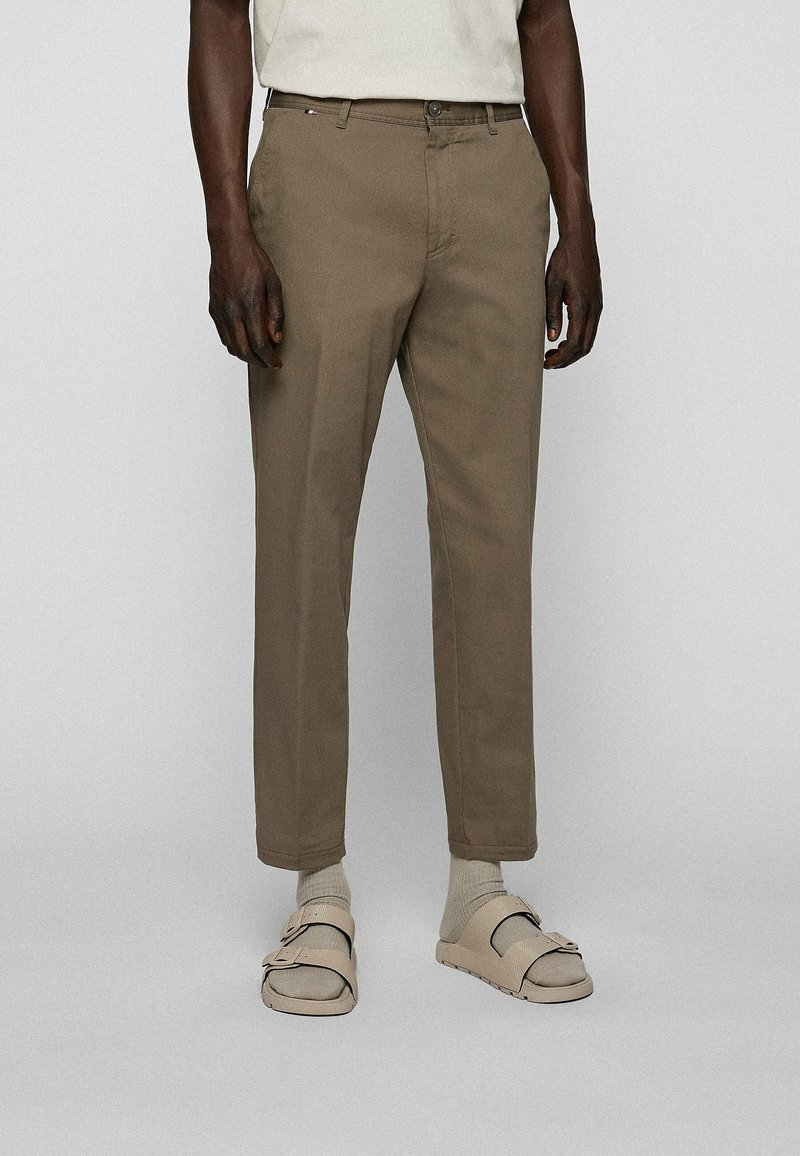 Homme portant un pantalon droit marron, des chaussettes beiges et des sandales beiges avec des boucles, debout devant un fond gris clair uni.