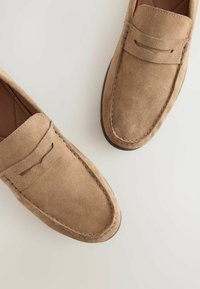 Vaaleat nubuk loafersit, joissa on sileä pinta, pyöreä kärki, ompeluyksityiskohtia ja koristeellinen liukstrap. Pohja on musta ja tasainen.
