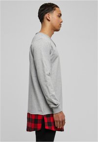 T-shirt à manches longues gris avec un ourlet à carreaux rouge et noir, arborant un col rond et des coutures visibles. Le matériau semble doux et texturé.