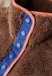 Tissu en fausse fourrure marron avec une texture douce, présentant une patte de boutonnage bleue et un biais de couleur corail. Les boutons sont étiquetés "Superdry".