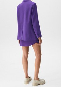 Blazer violet à coupe ajustée, avec des revers crantés et trois boutons. Associé à une jupe courte et des chaussures à semelles épaisses beiges.