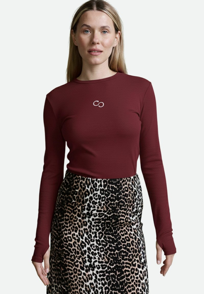 Bordeauxfarbener Langarmoberteil mit gerippter Textur und Unendlichkeitssymbol-Emblem; kombiniert mit einem Leopardenmusterrock in Beige-, Schwarz- und Brauntönen.