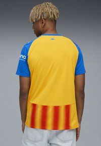 Camiseta de manga corta con una sección superior azul y una sección inferior amarilla con un patrón en degradado que transiciona a rojo. Diseño sin costuras.
