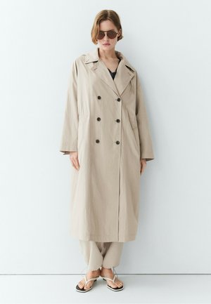 Femme portant un long trench-coat beige, un pantalon ample assorti, des lunettes de soleil et des sandales blanches, debout contre un mur blanc.