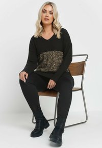 Mustapaita, jossa on leopardikuvioinen paneeli vyötäröllä, yhdistettynä mustiin legginsseihin ja mustiin nilkkureihin, istuu puutuolilla.