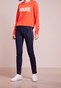 Pomarańczowa bluza z logo "HUGO", zestawiona z ciemnoniebieskimi jeansami z bocznymi pasami. Model ma na sobie białe trampki; widoczne gładkie faktury.