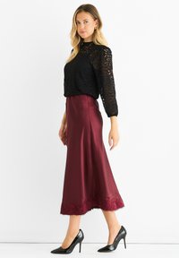 Woman wearing a black lace long-sleeve top with a burgundy satin midi skirt and black patent leather high heels, standing sideways.