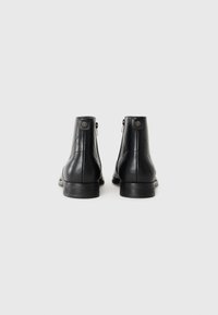 Par de botines de cuero negro con tacones bajos, cremalleras traseras visibles y pestañas decorativas en un fondo blanco liso.