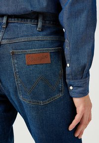 Jeans in denim con un patch in pelle marrone con il logo "Wrangler", cuciture gialle a contrasto e un design distintivo delle tasche a zigzag.