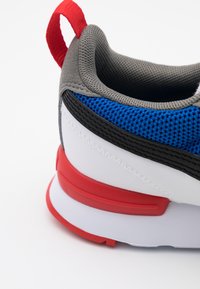 Tył sportowego buta z szarym materiałowym kołnierzem, niebieską dzianiną, czarnymi wykończeniami, białą cholewką oraz czerwonym uchwytem i detalem na podeszwie.