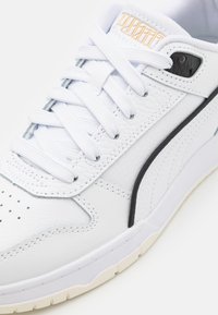 Hvide lædersneakers med tekstureret overflade, sort accentlinje og perforeret tå. Udstyret med hvide snørebånd og et logo i orange på tungen.