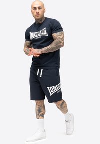 Navy bomulds t-shirt med hvid "Lonsdale London" tekst; kombineret med navy shorts med et lignende logo, suppleret med hvide sportsko.