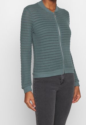 Teal textured zip-up bomber jacket with a ribbed collar and cuffs, featuring horizontal stripe patterns. Worn over dark gray jeans.