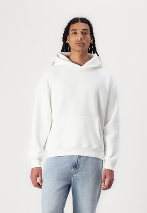 White hoodie made of soft fabric, featuring a front pocket, drawstring hood, and ribbed cuffs. Paired with light blue jeans.