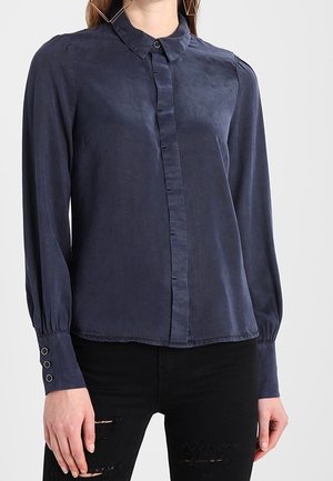 Kvinde iført en mørkeblå langærmet bluse med knapper og puffede manchetter, kombineret med sorte revnede jeans mod en ensfarvet baggrund.