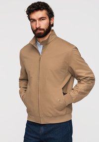 Homme barbu aux cheveux bouclés portant une veste zippée beige sur une chemise rayée, les mains dans les poches, debout devant un fond uni.