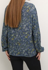 Chemise bleue à fleurs, à manches longues, avec un tissu texturé et une coupe ample. Fleurs dans des tons de jaune et de violet sur un fond bleu.