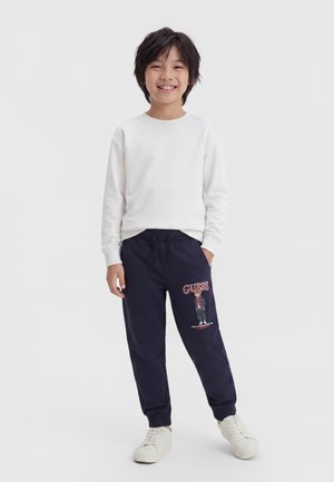 Enfant souriant, debout, portant un t-shirt blanc à manches longues, un pantalon de jogging bleu marine avec un motif d'ours et le texte "GUESS", ainsi que des baskets blanches.