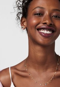 Collana di catena d'oro con pietre preziose multicolori e piccoli ciondoli; il modello ha i capelli ricci e un incarnato chiaro, indossa orecchini a goccia.