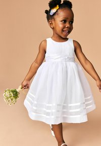 Robe blanche sans manches avec un corsage ajusté, jupe ample, rayures horizontales au bas, fabriquée en tissu léger, accompagnée de chaussures blanches.
